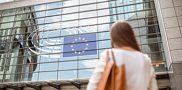 Foto einer Frau, die auf ein europ&auml;isches Regierungsgeb&auml;ude mit der Europaflagge im Fenster blickt
