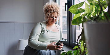 Eine Frau sitzt am Fenster in einer Wohnung und schaut l&auml;chelnd auf ihr Smartphone. Tageslicht f&auml;llt durch das Fenster, daneben steht eine gr&uuml;ne Zimmerpflanze im Vordergrund.