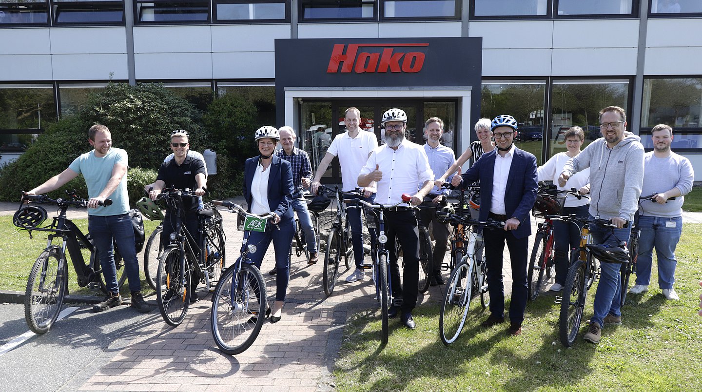 Foto zeigt die Eröffnung der landesweiten Mitmachaktion. Eine Gruppe Menschen steht mit Fahrrädern vor dem Firmensitz Hako GmbH. Alle Personen sehen gut gelaunt aus.