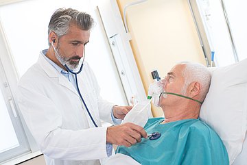 Rauchen und Krankenhauseinweisungen in Westfalen-Lippe Ein älterer Patient liegt im Krankenhaus im Bett. Er trägt eine Sauerstoffmaske und wird gerade von einem Arzt mit Stethoskop auf der Brust abgehorcht.