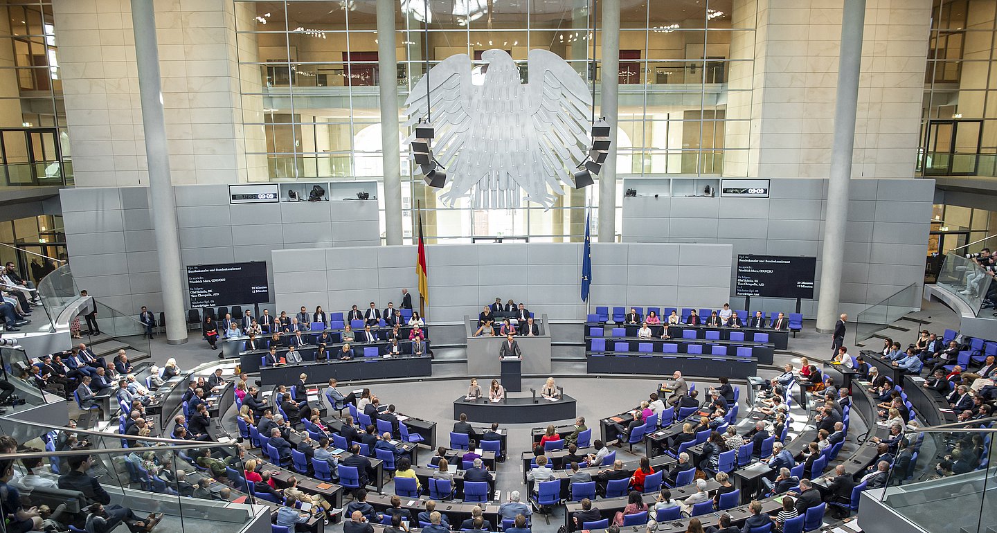 Das Bild zeigt eine Totale vom Plenum des Bundestag. Zentral in der oberen Bildhälfte sieht man den Bundesadler, darunter Präsidium, Rednerpult und ein volles Plenum