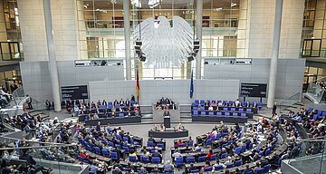 Das Bild zeigt eine Totale vom Plenum des Bundestag. Zentral in der oberen Bildh&auml;lfte sieht man den Bundesadler, darunter Pr&auml;sidium, Rednerpult und ein volles Plenum
