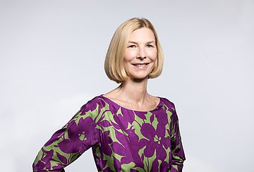 Dr. Isabella Erb-Herrmann Das Porträtbild zeigt eine blonde Frau mit gescheiteltem Haaren bis zum Kinn und violett-grün gemusterter Bluse ohne Kragen.