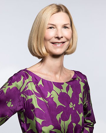 Dr. Isabella Erb-Herrmann Das Porträtbild zeigt eine blonde Frau mit gescheiteltem Haaren bis zum Kinn und violett-grün gemusterter Bluse ohne Kragen.