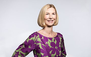 Dr. Isabella Erb-Herrmann Das Porträtbild zeigt eine blonde Frau mit gescheiteltem Haaren bis zum Kinn und violett-grün gemusterter Bluse ohne Kragen.