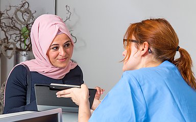 Foto: Eine ärztliche Assistentin reicht einer Frau, die ein Kopftuch trägt, ein Klemmbrett mit Formularen.
