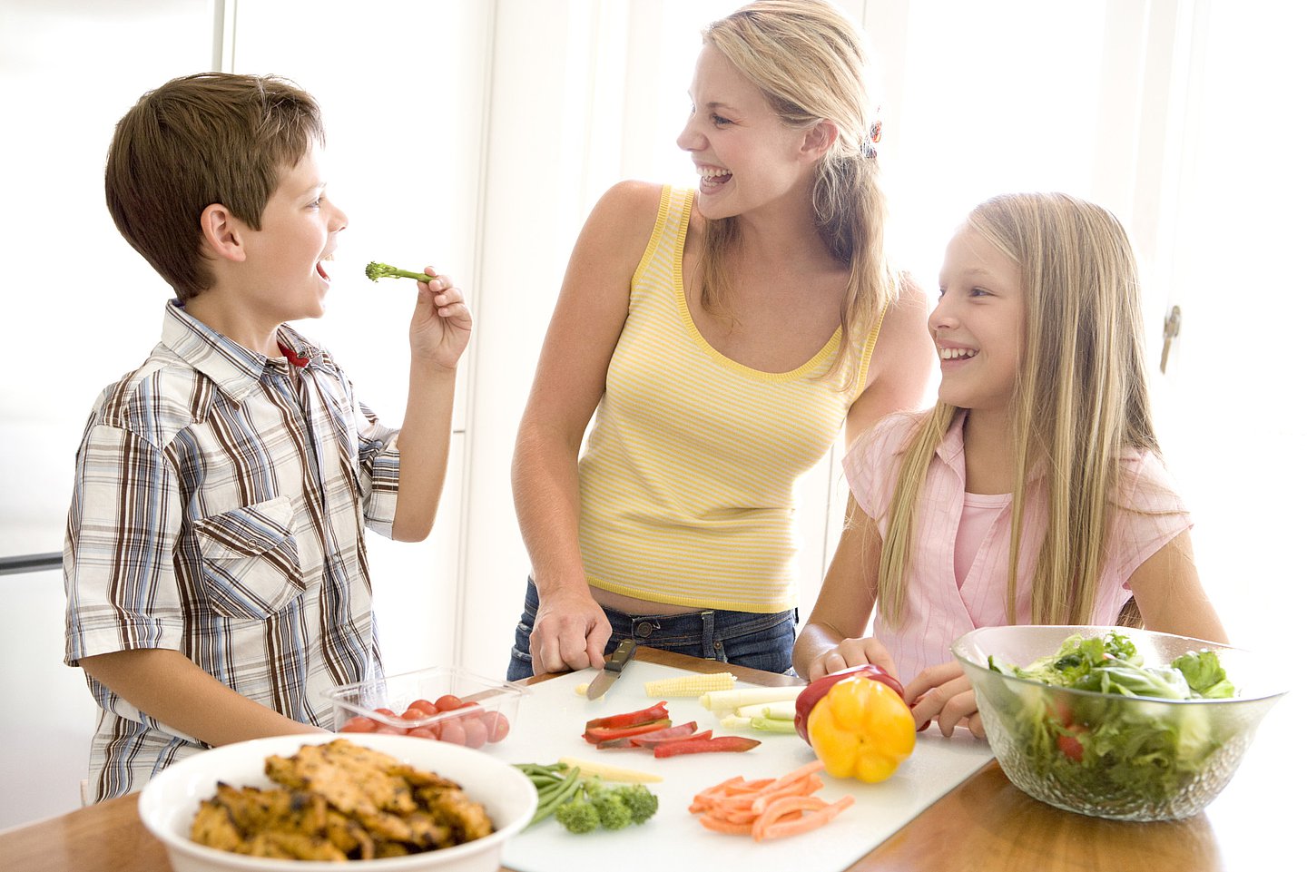 Eine Mutter steht mit ihren Kindern in der Küche. Auf einem Schneidebrett vor ihnen liegt geschnittenes Gemüse, daneben steht ein Salatschüssel. Der Junge steckt sich gerade ein Stück Rohkost in den Mund.