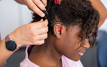 Foto einer jungen Frau, der hinter dem Ohr die Sendespule f&uuml;r ein Cochlea-Implantat angebracht wird
