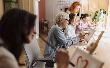 An einem langen Tisch sitzen verschiedene Personen und bemalen eine Leinwand.