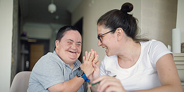 &Auml;ltere Frau mit Down-Syndrom, die fr&ouml;hlich eine j&uuml;ngere Frau abklatscht. Beide sitzen in einer Einrichtung.