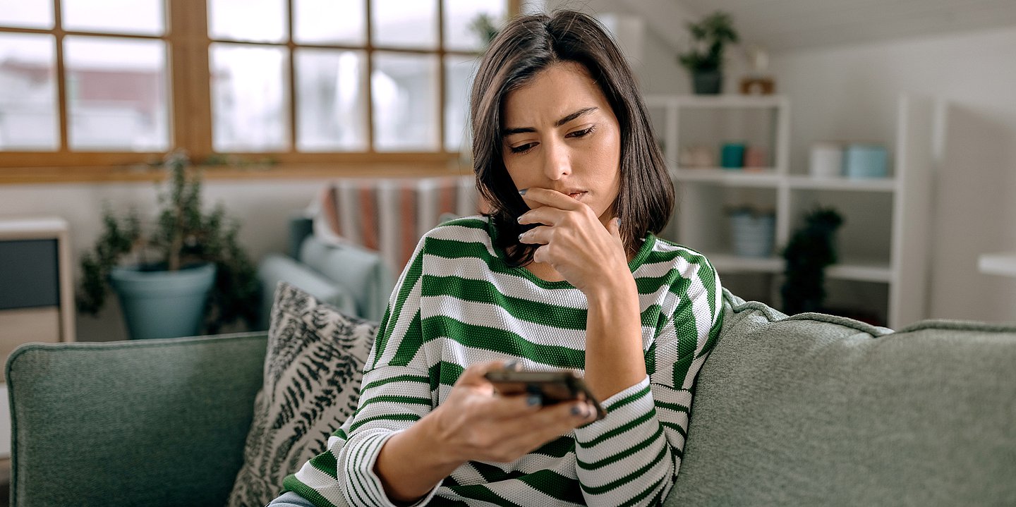EIne Frau sitzte auf dem Sofa und schaut auf das Handy in ihrer Hand, sie schaut besorgt und hat die andere Hand am Mund.