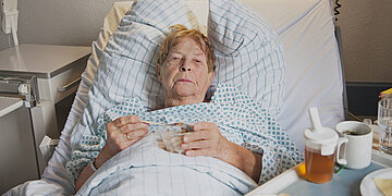 Eine &auml;ltere Dame liegt im Krankenhausbett mit einer Schale Essen auf ihrem Bauch. Sie schaut abwesend und m&uuml;de drein.