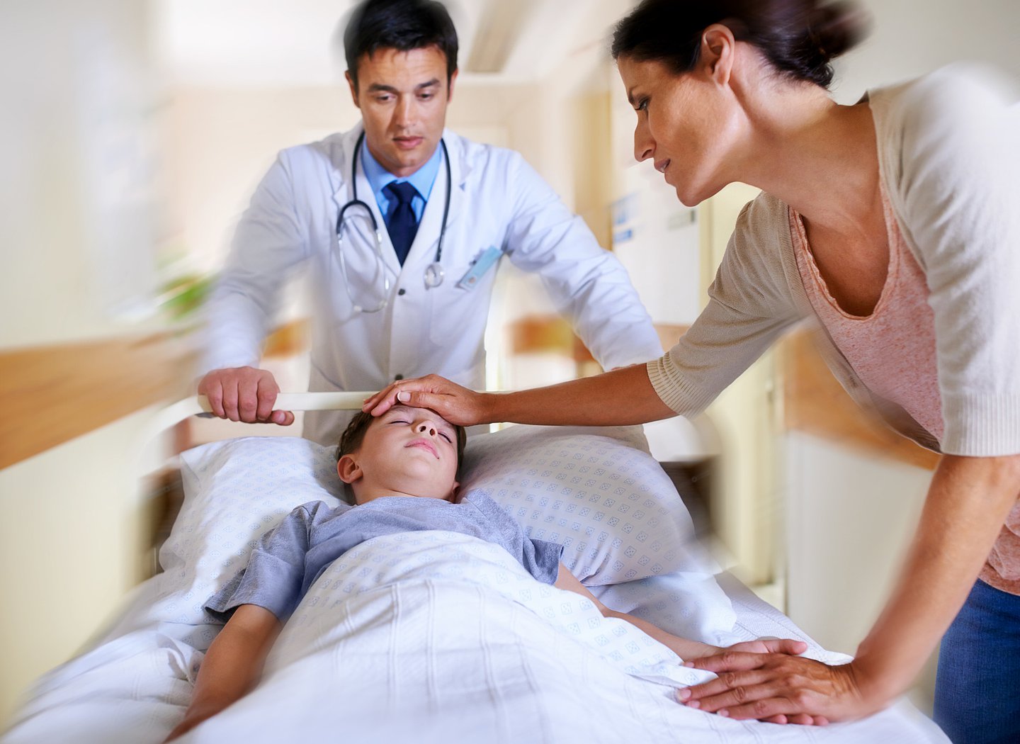 Ein Kind wird auf einer Trage über einen Krankenhausflur geschoben, vorweg ein Arzt, an der Seite geht die Mutter mit.