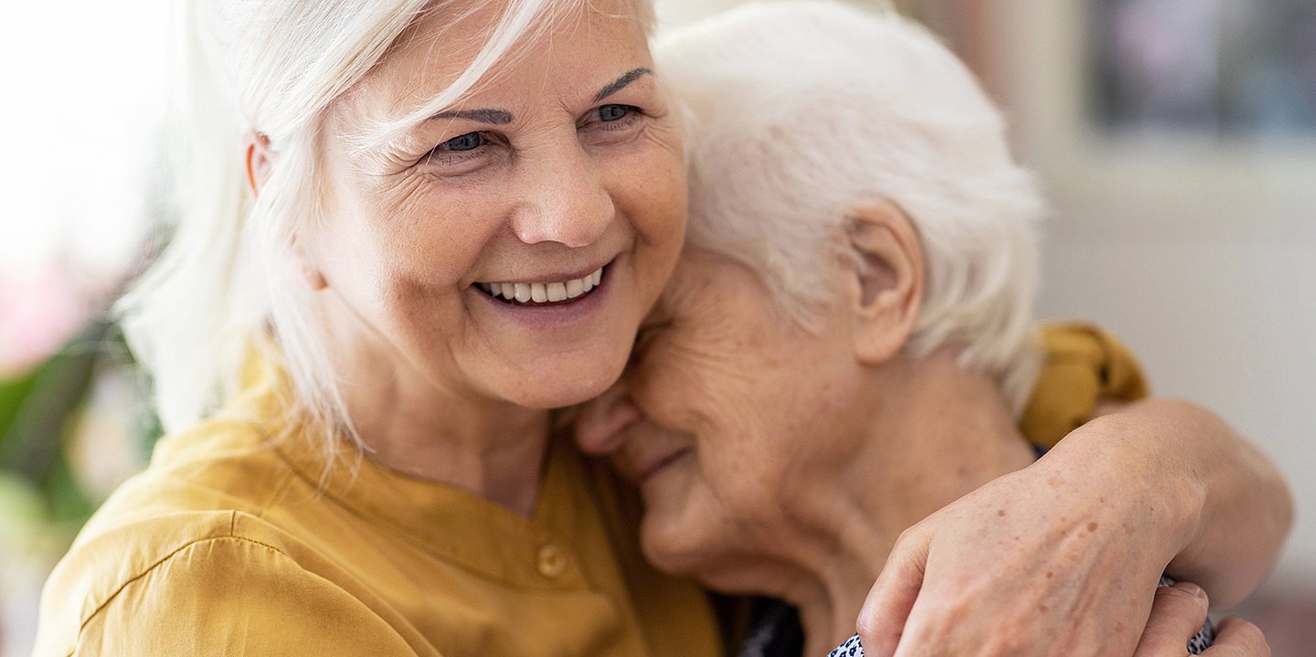 Foto: Eine jüngere Frau nimmt eine ältere Frau in den Arm und lächelt.