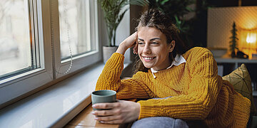 Foto einer jungen Frau, die sich gl&uuml;cklich grinsend mit einer Tasse Tee in der Hand auf ein Fensterbrett st&uuml;tzt und nach drau&szlig;en blickt