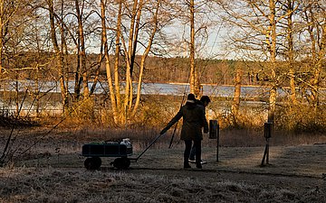 Zwei Personen mit einem Bollerwagen am winterlichen Ufer des Stechlinsees bei zwei kleinen Messstationen zur Messung der Luftqualität