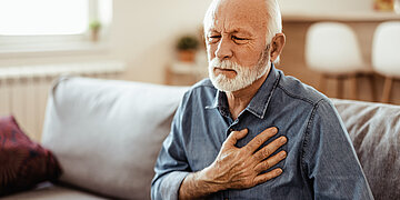Ein &auml;lterer Herr sitzt mit schmerzverzerrtem Gesicht auf der Couch und greift sich an die Brust. 