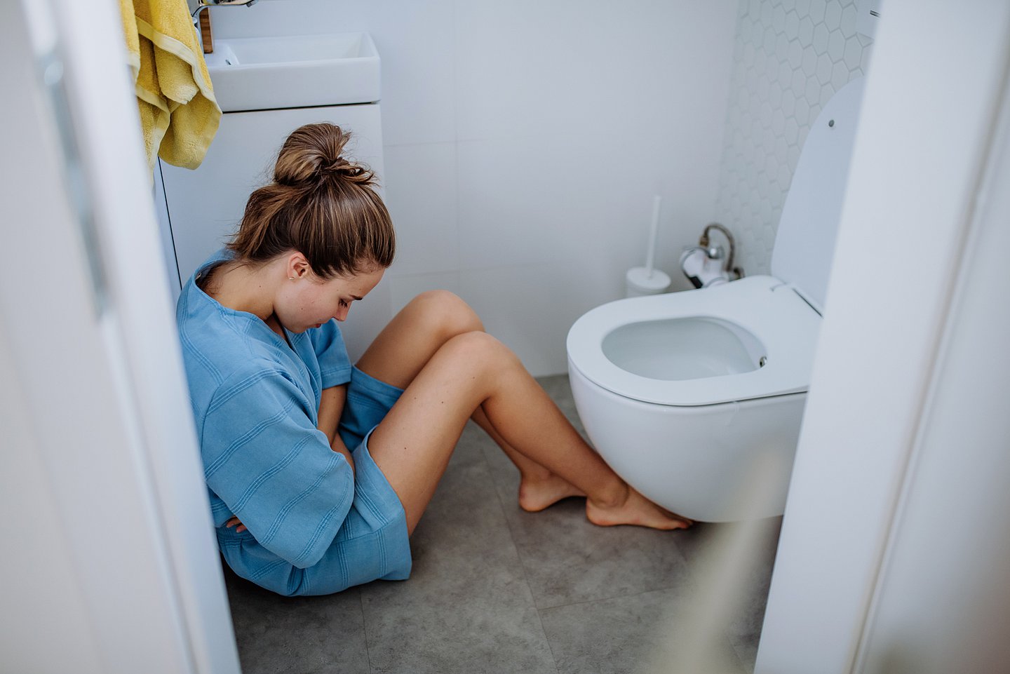 Foto zeigt eine Frau, die in einem WC auf dem Boden vor der Toilette sitzt und sich den Bauch hält.