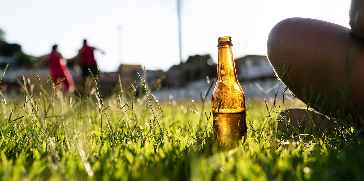 Auf einer Wiese spielen Kinder im Hintergrund Fußball. Im Vordergrund sitzt ein Mensch, neben ihm steht eine Bierflasche im Gras.