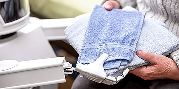 Foto: Eine &auml;ltere Person hat Handt&uuml;cher in der Hand und ein Roboter h&auml;lt ebenfalls die Handt&uuml;cher