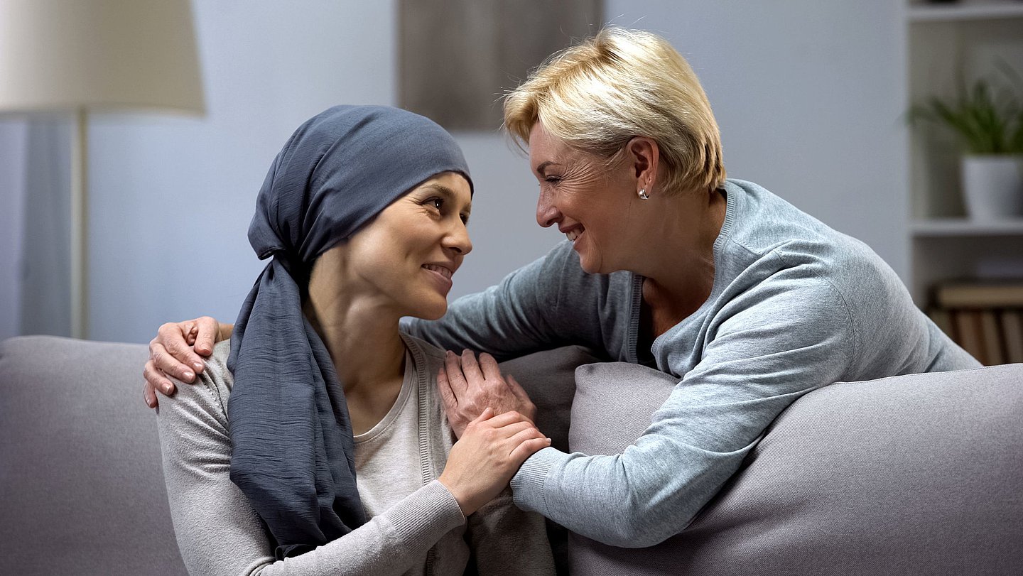 Foto einer kranken Frau, die auf dem Sofa sitzt und Zuwendung von einer Hospizmitarbeiterin erhält, die ihr den Arm um die Schulter legt..