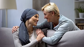 Fördermittel für ambulante Hospizdienste Foto einer kranken Frau, die auf dem Sofa sitzt und Zuwendung von einer Hospizmitarbeiterin erhält, die ihr den Arm um die Schulter legt..