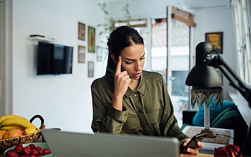 Eine Person sitzt am Laptop und schaut konzentriert auf ihr Smartphone.