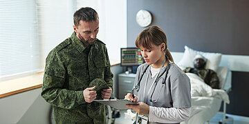 In einem Krankenhauszimmer stehen zwei Personen nebeneinander: Eine &Auml;rztin mit Klemmbrett und ein Bundeswehrsoldat in Uniform. Im Hintergrund liegt ein verwundeter Soldat mit Kopfverband im Krankenhausbett.