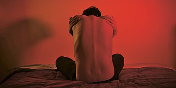 Rückenschmerzen Foto eines Mannes, der mit dem Rücken zur Kamera im Schneidersitz da sitzt. Er hat einen nackten Oberkörper und umarmt sich mit beiden Händen, als hätte er Schmerzen.
