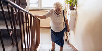 Herzschwäche Foto: Eine ältere Dame, läuft Treppen, hält sich am Geländer fest und sieht angestrengt aus.