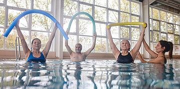 &Auml;ltere Menschen machen Wassergymnastik in einem Schwimmbecken mit Schaumstoffnudeln, angeleitet von einer Trainerin.