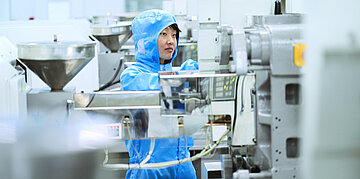 Eine Arbeiterin in blauer Schutzkleidung und Haube bedient Maschinen in einer sauberen industriellen Produktionsumgebung.