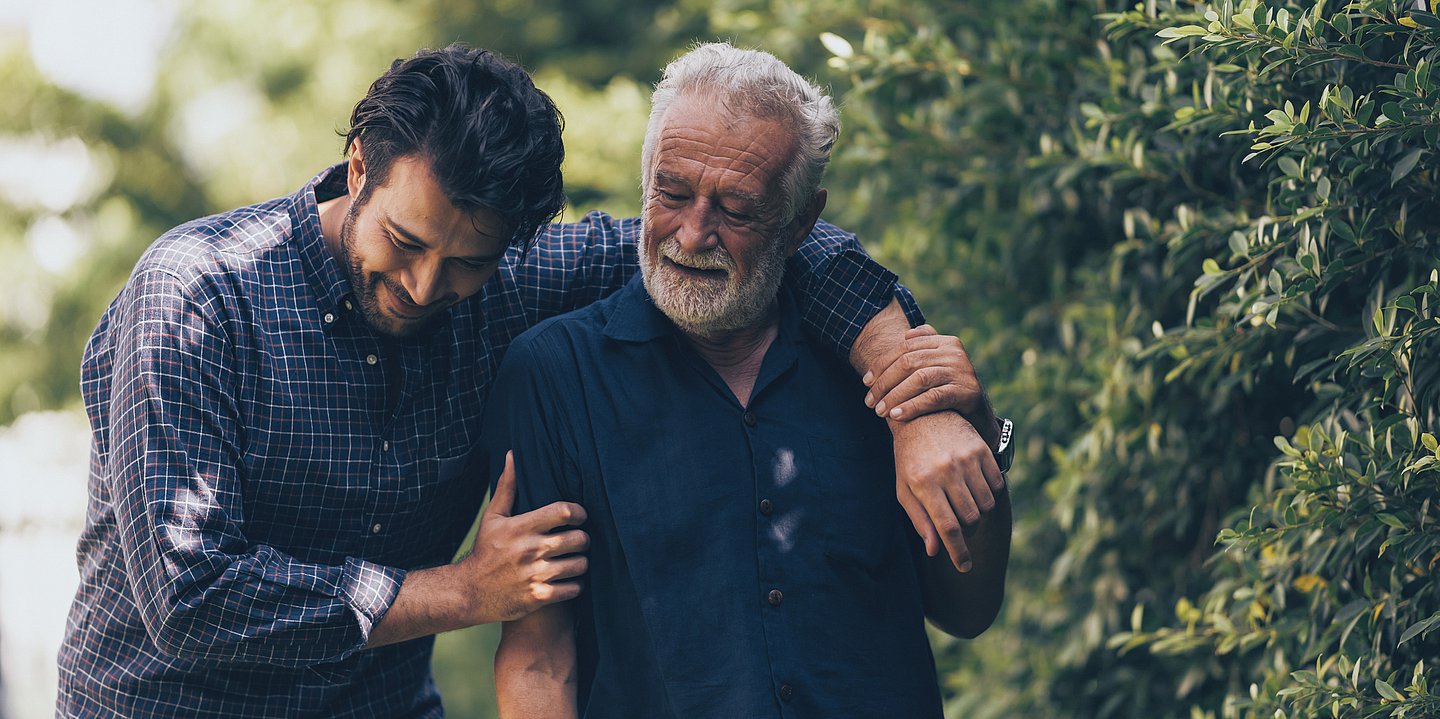 Foto: Ein junger Mann und ein älterer Mann gehen spazieren, währen der junge Mann seinen Arm um den Älteren legt.