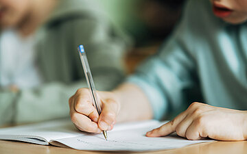 Foto: Ein Sch&uuml;ler sitzt an einem Tisch und schreibt in ein Heft. 