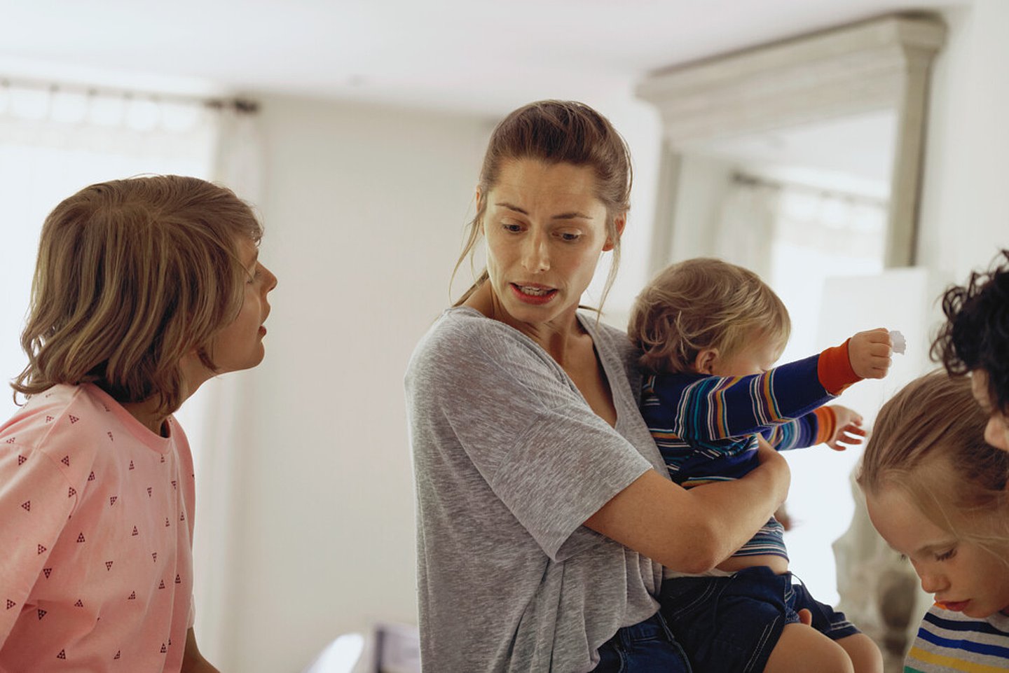 Symbolbild für Stress einer Alleinerziehenden: Eine gestresste Mutter hält ein kleines Kind im Arm und diskutiert mit einem Jugendlichen, während weitere jüngere Kinder am Bildrand zu sehen sind.