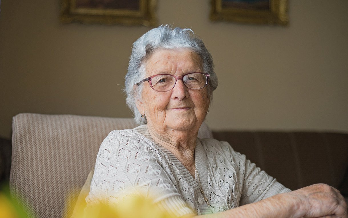 Foto einer alten Frau, die auf einem Sofa sitzt und lächelnd in die Kamera blickt