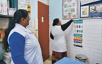 In einem Praxiszimmer stehen zwei Frauen. Die eine zeigt auf ein Plakat an der Wand.