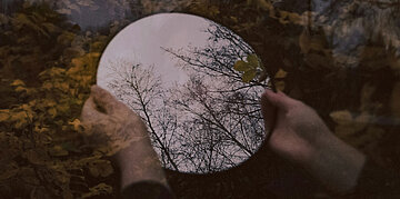 Bl&auml;tterhintergrund: Davor halten zwei H&auml;nde einen runden Spiegel.