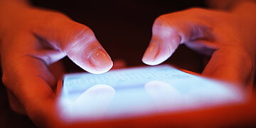 Foto: Nahaufnahme eines Smartphones mit roter H&uuml;lle, das von zwei H&auml;nden gehalten wird. Die Daumen tippen auf der Tastatur.
