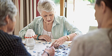 Eine Frau puzzelt an einem Tisch, zwei andere schauen ihr dabei zu.