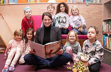 AOK-Landesdirektorin Iris Kröner sitzt mit acht Kita-Kindern zusammen und liest aus einem Buch vor. 