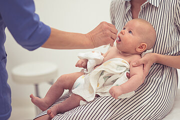 Eine junge Mutter hat ihren S&auml;ugling im &auml;rztlichen Behandlungszimmer auf dem Scho&szlig;, der von einem Arzt/einer &Auml;rztin eine L&ouml;sung in den Mund verabreicht bekommt.