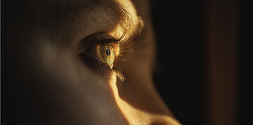 ahaufnahme eines menschlichen Auges im Profil, das von warmem, goldenem Licht beleuchtet wird; der Hintergrund ist dunkel.