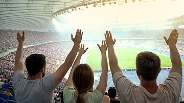 AOK-Umfrage Sport-Events in Westfalen-Lippe Foto zeigt ein volles Fußballstadion mit Blick auf das Spielfeld und jubelnde Fans, die die Arme hochreißen.