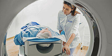 Eine medizinische Fachkraft schiebt einen &auml;lteren Patienten auf einer Liege in ein MRT- oder CT-Ger&auml;t zur Untersuchung.