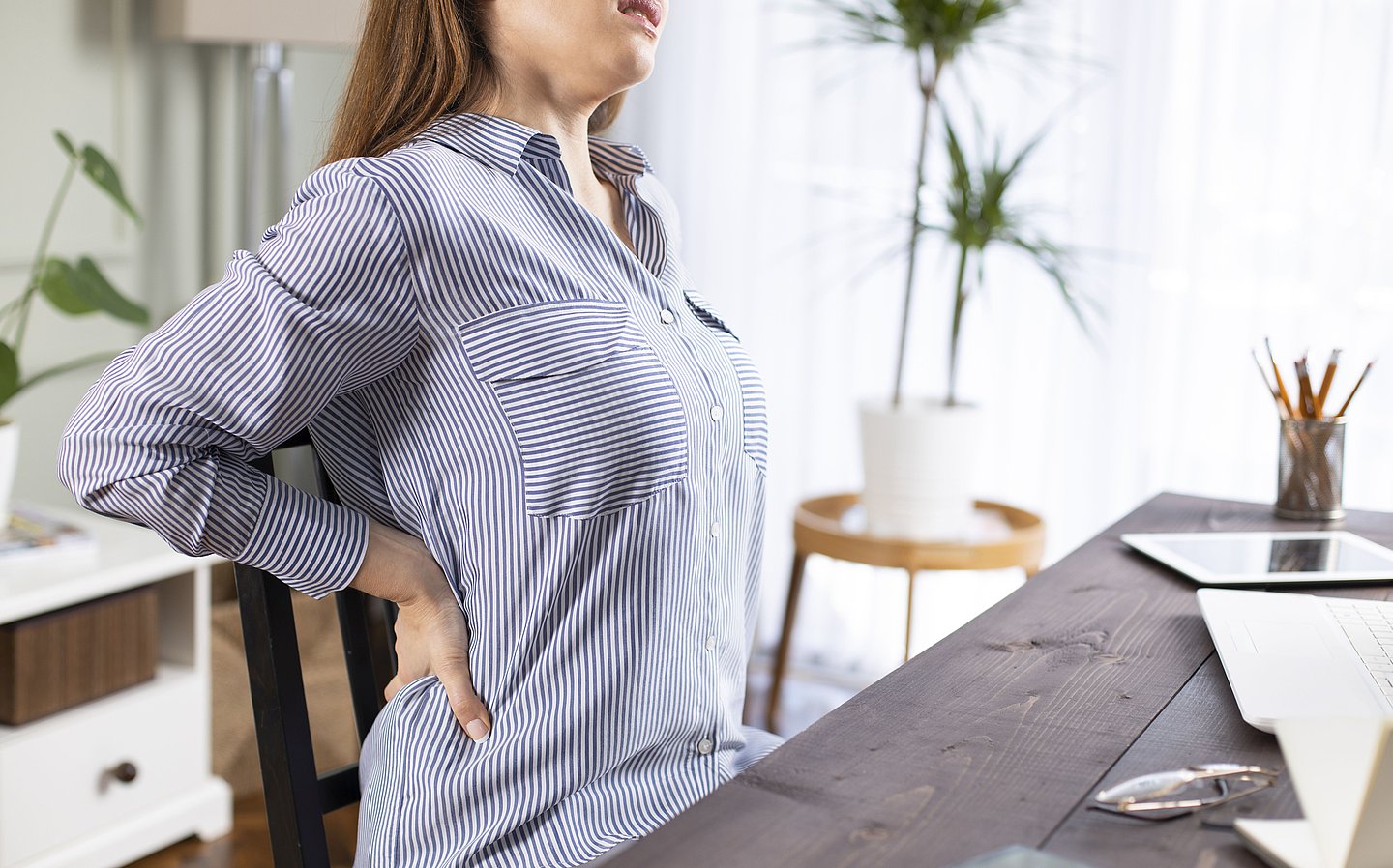 Foto: Eine Frau sitzt am Tisch und fasst sich an ihren schmerzenden Rücken.