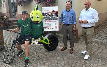 Drei Personen stehen vor einem Geb&auml;ude in Haslach und zeigen das Plakat des Firmenlaufs 2026.