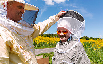 Zwei M&auml;nner, darunter der Bewohner eines Pflegebauernhofs, stehen mit Schutzkleidung gegen Bienen auf einem Feld.