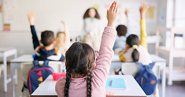 Der Blick aus Sch&uuml;lerperspektive nach vorn zur Lehrerin in einem Klassenzimmer. Man sieht die R&uuml;cken der sitzenden Kinder, von denen einige sich melden.