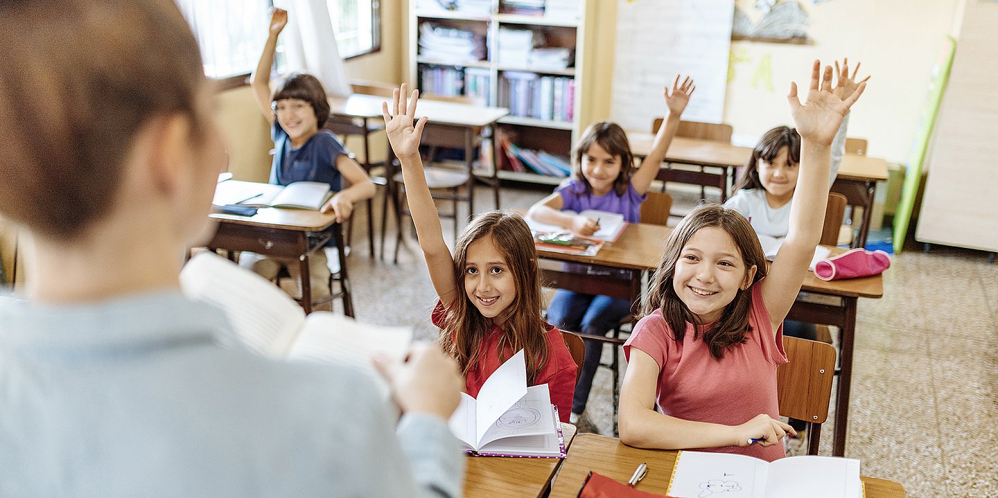 Foto: Eine Schulklasse sitzt im Unterricht und meldet sich. Vorne steht eine Lehrerin.